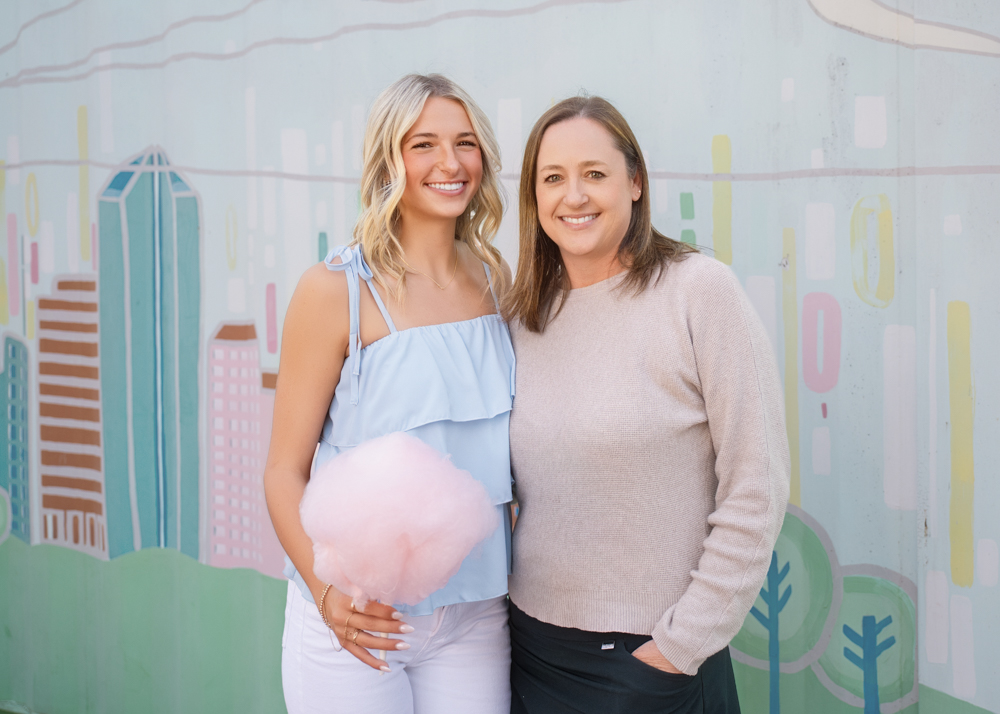 Two women smiling in front of a pastel mural depicting city buildings. One holds pink cotton candy. The mood is cheerful and relaxed.