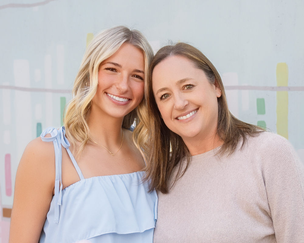 Senior girl and her mom smiling warmly, standing close together against a soft pastel background. One wears a light blue dress, the other a beige sweater, conveying a sense of joy and connection.