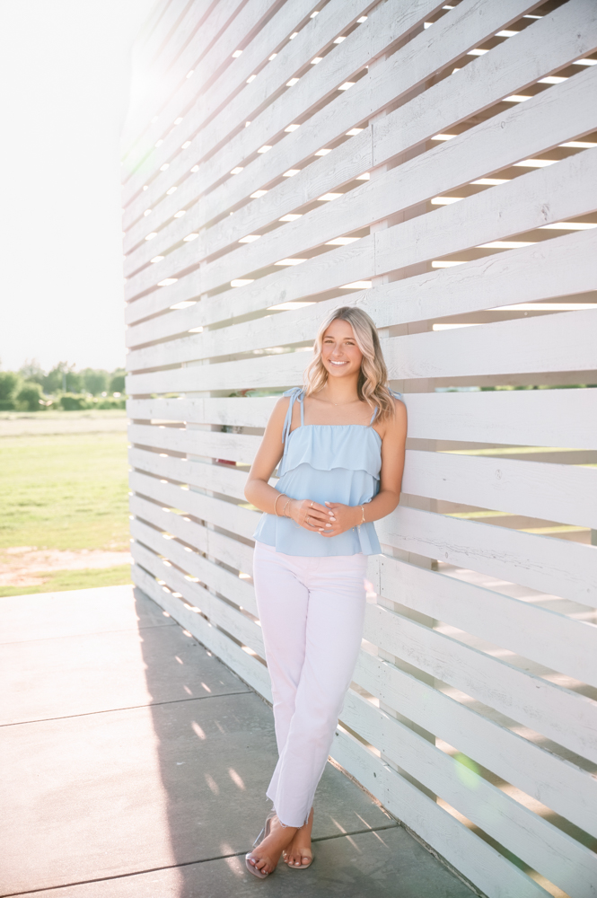 A woman in a light blue top and white pants leans against a white slatted wall, smiling softly. Sunlight filters through the slats, creating a serene, summery vibe.
