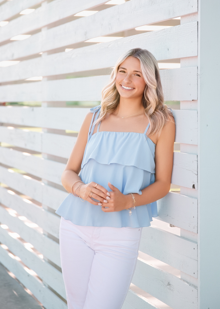 Smiling woman with wavy hair in a blue top and white pants leans on a light wooden wall. The scene is bright and airy, exuding a cheerful vibe.