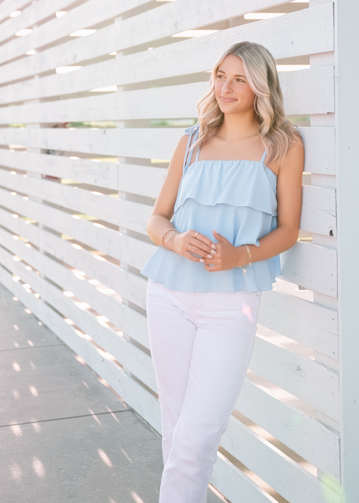 Young woman in a light blue ruffled blouse and white pants leans on a white slatted wall, smiling softly. Soft lighting creates a calm, serene mood.