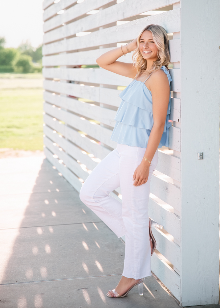 A woman in a light blue top and white pants leans against a slatted white fence. Sunlight casts stripes of shadow. She smiles, exuding a relaxed, cheerful vibe.