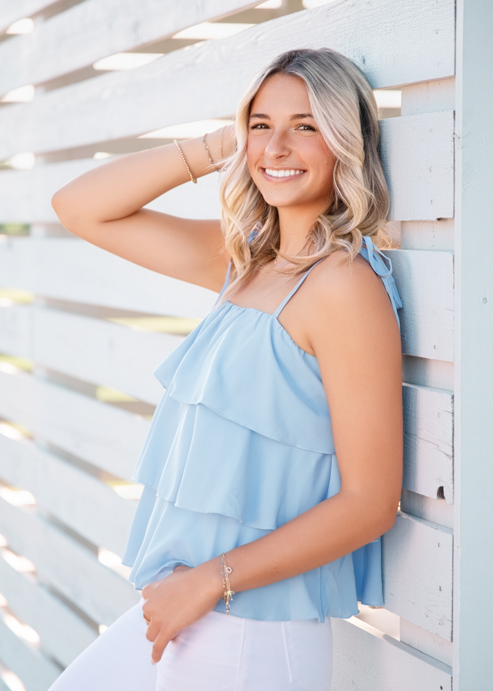 Smiling person with wavy blonde hair wearing a light blue ruffled top and white jeans, leaning against a white wooden fence, exuding a cheerful vibe.