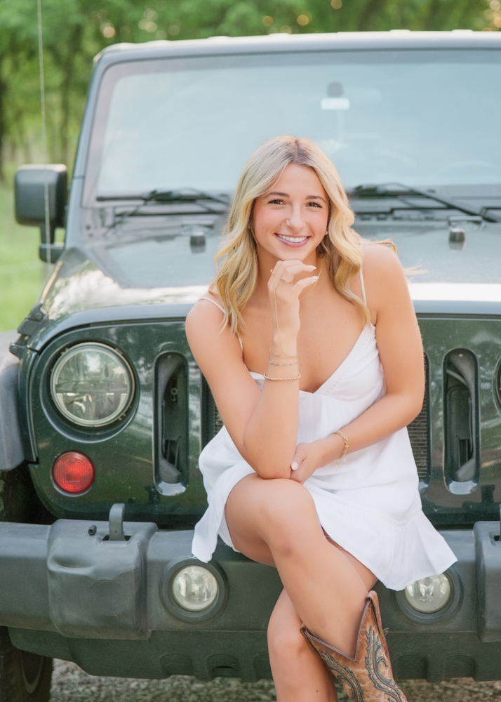 Young woman in a white dress and cowboy boots sits on a parked Jeep. She smiles warmly with a hand under her chin, against a green, sunlit backdrop.