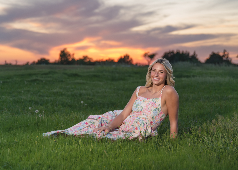 Kenna in a floral dress reclines on a lush green field, smiling warmly. The sunset in the background creates a serene and joyful ambiance.