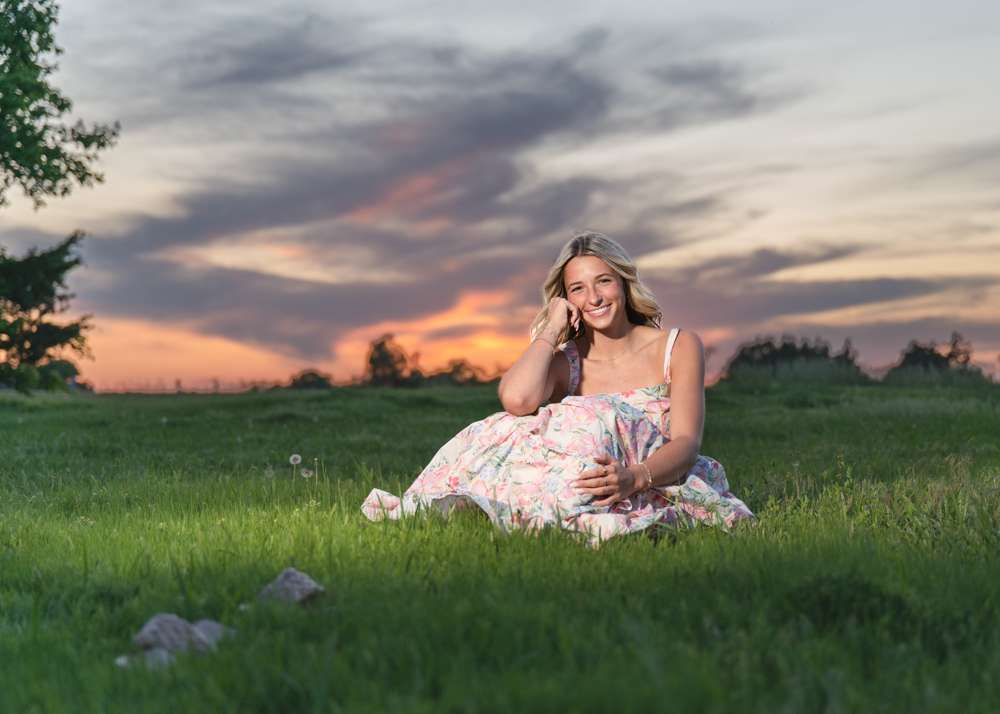 A woman in a floral dress sits on grass at sunset, smiling warmly. The sky is vivid orange and pink, creating a serene, joyful atmosphere. Trees are silhouetted in the background.