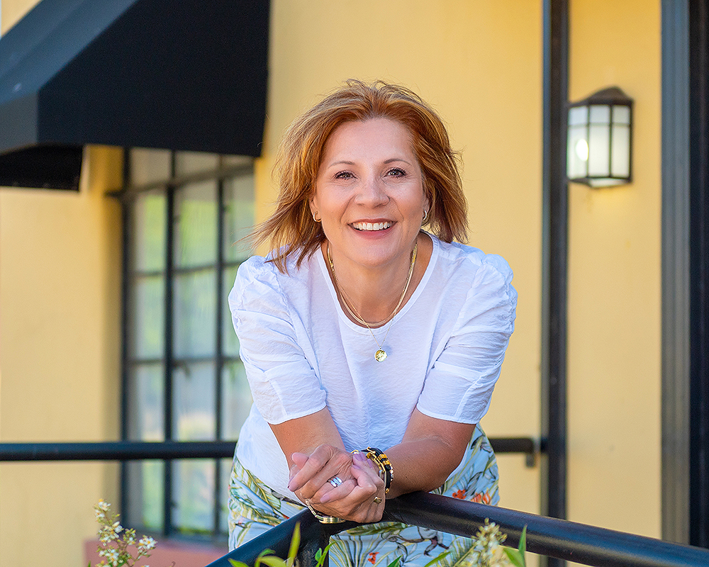 A woman in a white blouse leans on a black railing, smiling warmly. She's outside near a yellow wall with a lamp, conveying a cheerful atmosphere.