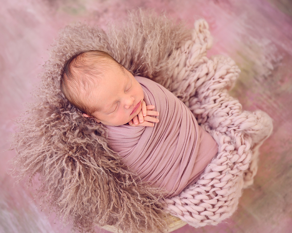 Newborn baby peacefully sleeping, wrapped in a soft, pink blanket, surrounded by textured fur on a pastel backdrop.