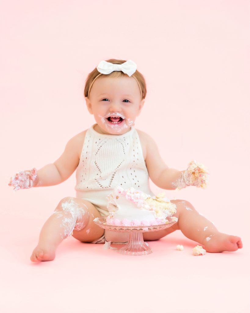 A joyful baby in a white outfit and headband sits on a pink background, laughing, with cake on their face and hands, beside a partially smashed cake.