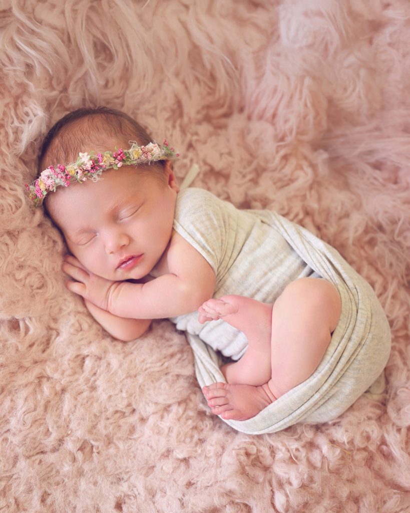 Sleeping newborn wrapped in a soft, beige blanket, lying on a fluffy pink surface. The baby wears a delicate floral headband