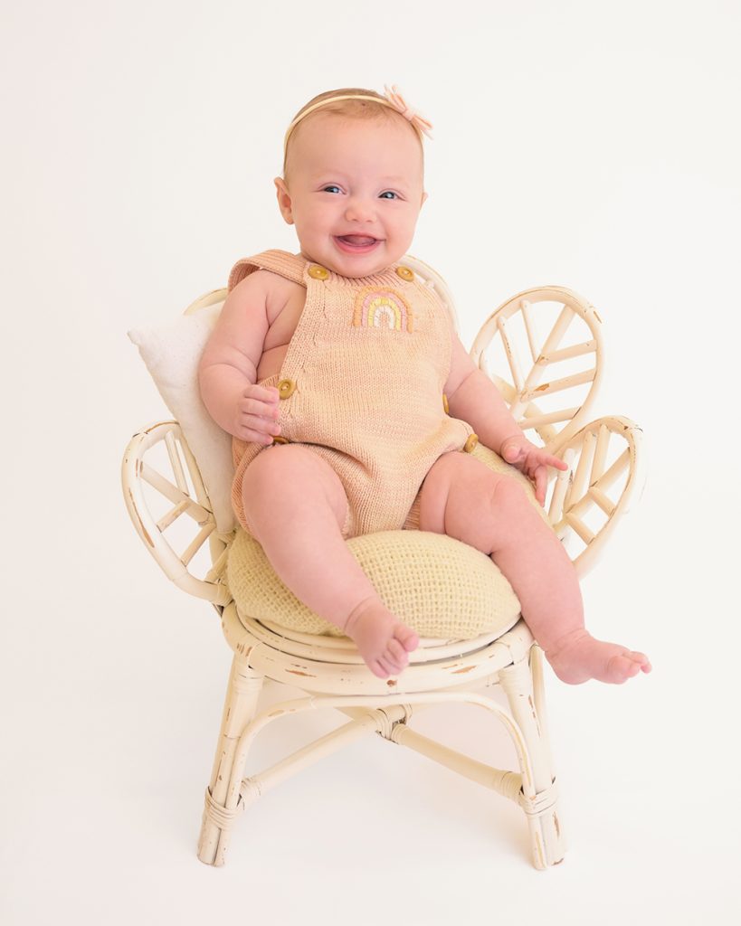 A smiling baby in a peach romper with a rainbow design sits on a small rattan chair.