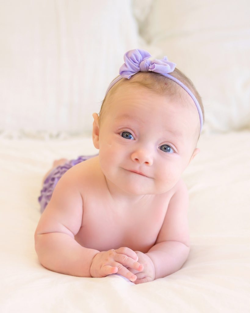 A baby lies on a soft, white blanket, wearing a purple headband with a bow.