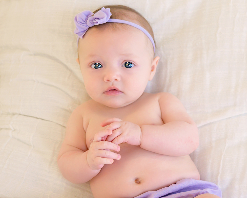 A smiling baby in a peach romper with a rainbow design sits on a small rattan chair.