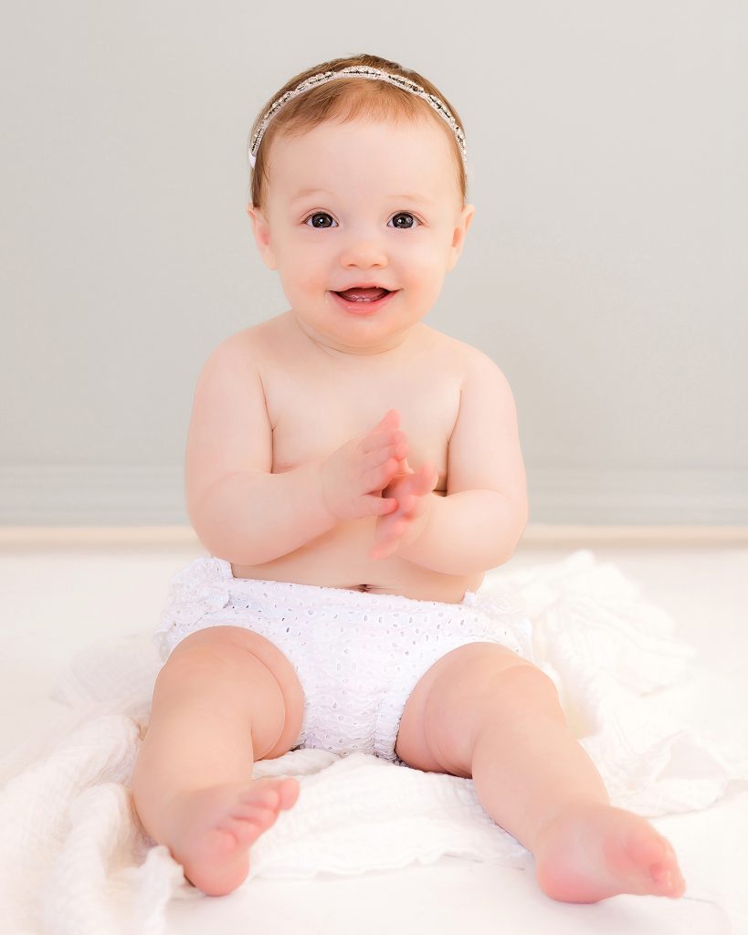 A joyful baby wearing a white headband and diaper sits on a soft white blanket, clapping hands and smiling against a light gray background.