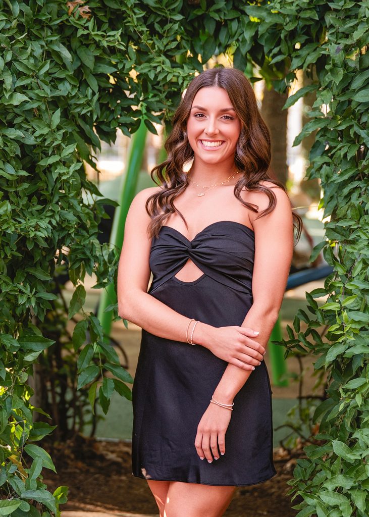 Smiling high school senior in a black dress stands in a lush, green leafy archway. Her arms are crossed gently, exuding confidence and elegance.