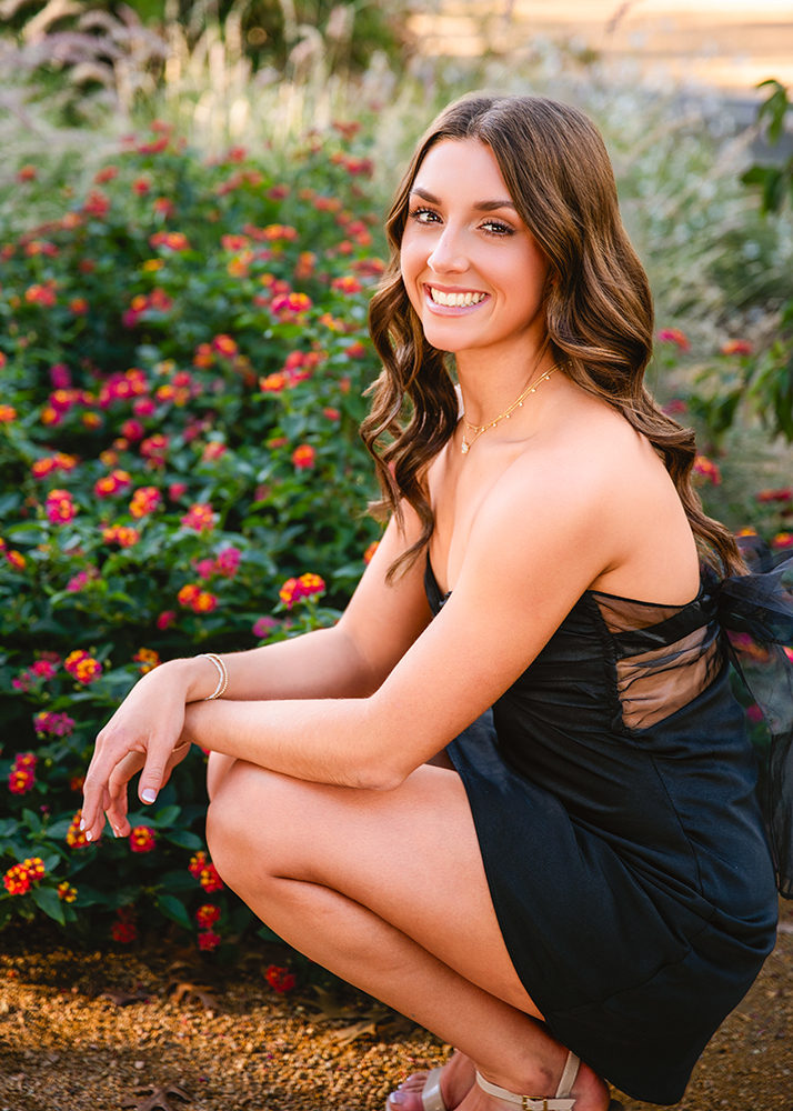A woman in a black dress crouches near vibrant flowers, smiling warmly. The scene is bright and colorful, conveying joy and serenity.