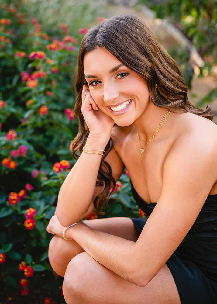 Macey poses outdoors for her senior photos, resting her head on her hand. She wears a black strapless top and gold jewelry. In the background, vibrant red and yellow flowers create a cheerful, sunny atmosphere.