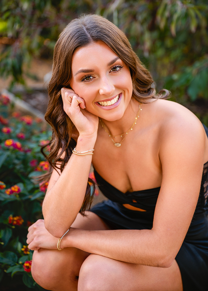 A woman in a black dress smiles warmly, resting her head on her hand. She is surrounded by greenery and red-orange flowers, creating a cheerful tone.