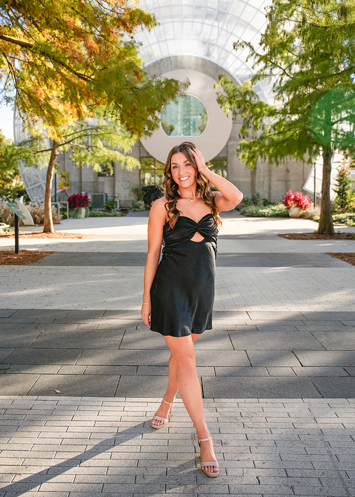 Macey's senior photos on a sunny pathway at the Myriad Botanical Gardens, smiling confidently with lush green trees and a modern glass building in the background.