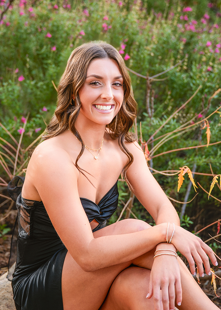 Senior girl poses for photos outdoors, surrounded by green foliage and pink flowers. The scene conveys a joyful and serene mood.