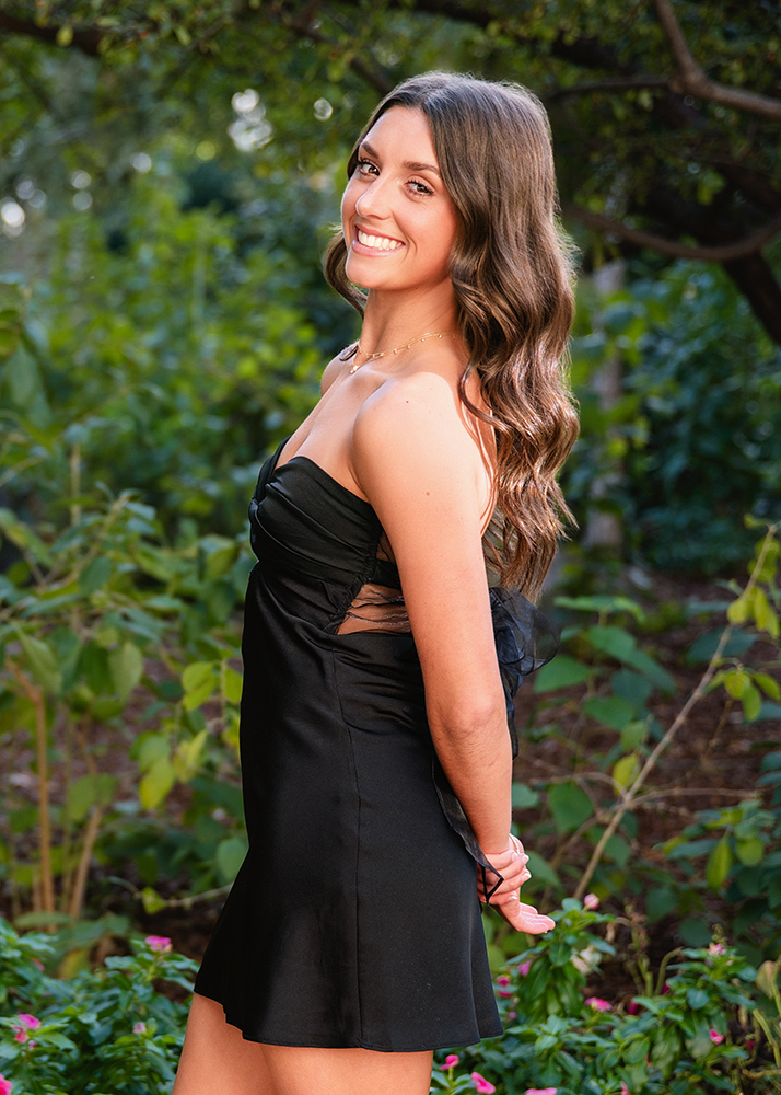 A senior in a black dress smiles warmly while standing in a lush garden. Her wavy hair is down, and she stands with her arms behind her back.
