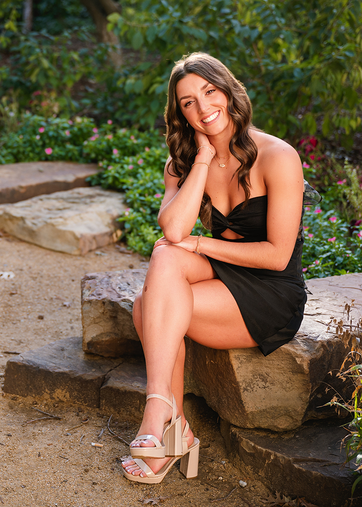 A senior girl in a black dress smiles warmly, seated on a stone step in a garden. She's surrounded by greenery and flowers, exuding a relaxed, cheerful vibe.