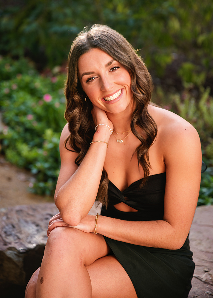 A high school senior girl in a black dress with long, wavy hair smiles warmly while seated in a lush garden setting. The mood is joyful and relaxed.
