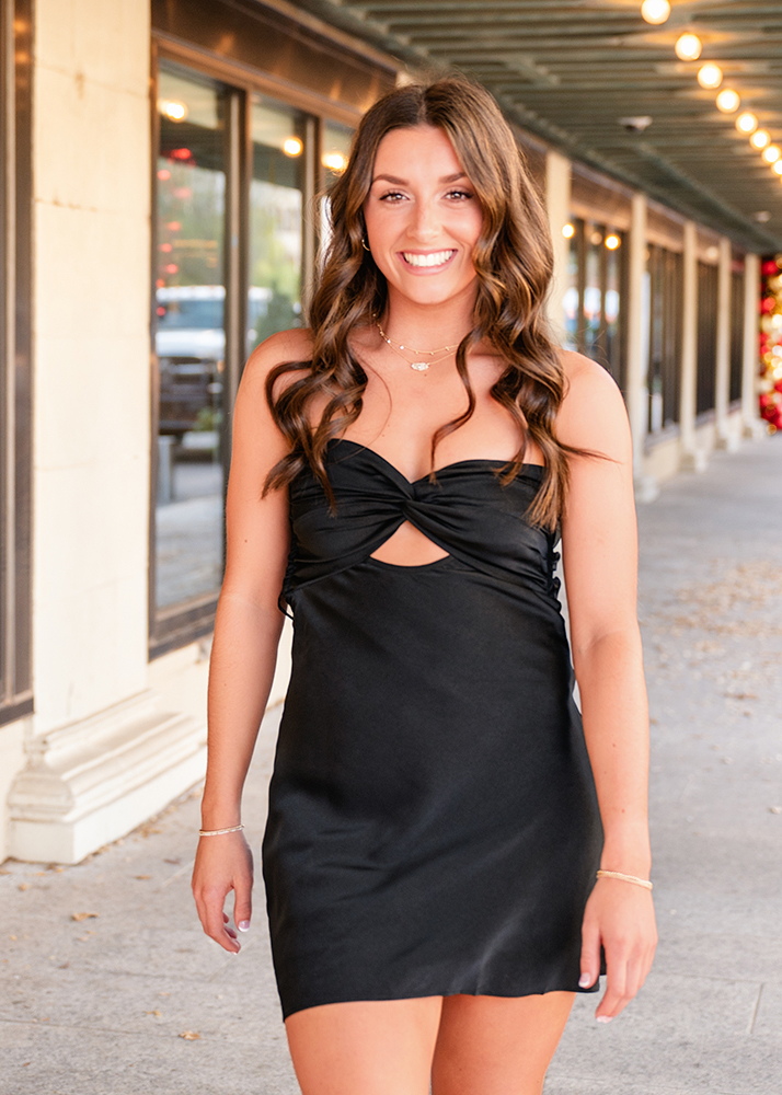 Young woman in a black dress smiles while walking on a sidewalk under a string of lights at an outdoor shopping area. Elegant and cheerful vibe.