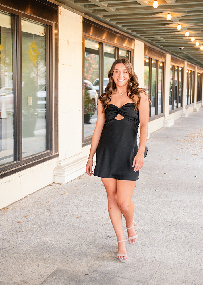 Macey walks down a sidewalk under string lights in downtown OKC, with glass windows and reflections beside her. The scene feels casual and upbeat.