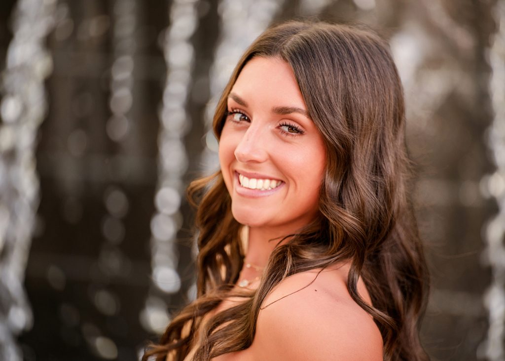 A senior girl with long, wavy brown hair smiles warmly against a blurred, sparkling waterfall background. The atmosphere is joyful and radiant.