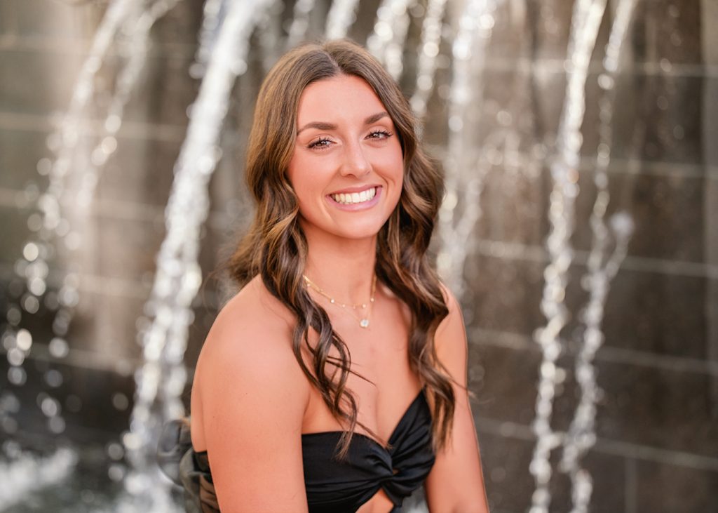 A high school senior girl with long, wavy brown hair smiles warmly against a blurred, sparkling waterfall background. The atmosphere is joyful and radiant.