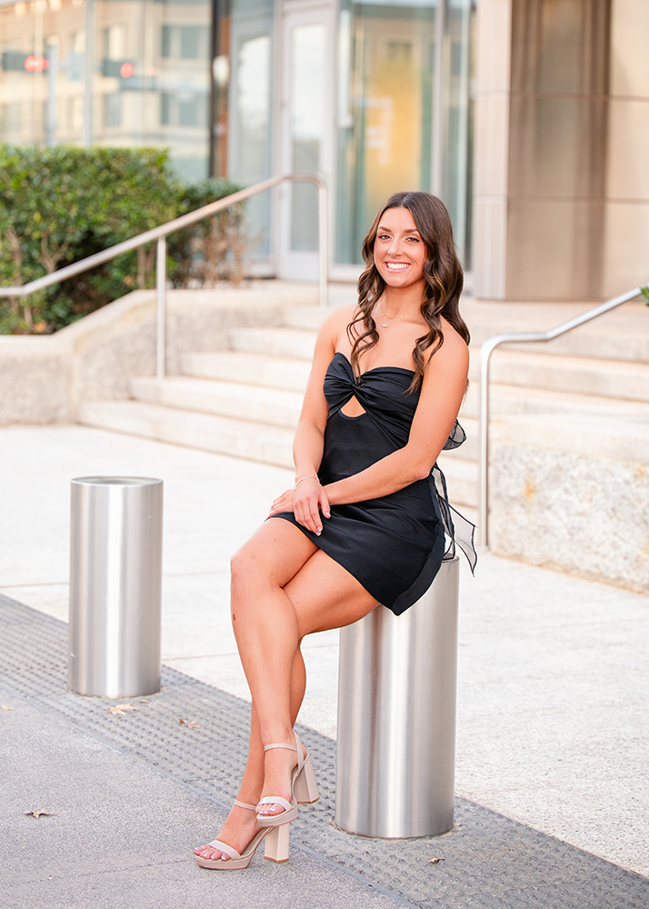 Macey sits on a metal cylinder in an urban setting in downtown Oklahoma City. Background includes stairs and bushes, conveying a cheerful, formal tone.