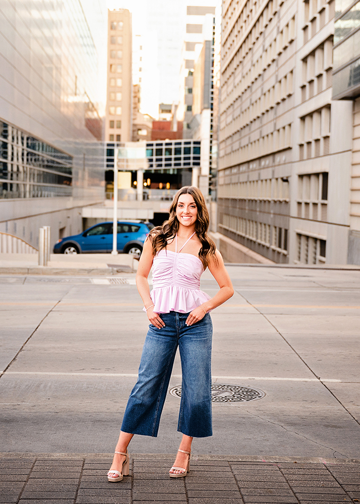 A high school senior stands confidently on an urban street, wearing a pink top and jeans. The background features modern buildings and a parked blue car.