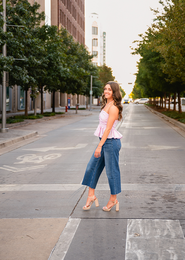 Senor girl in a pink top and denim pants smiling, walks across an empty street lined with trees, conveying a cheerful and serene urban vibe.