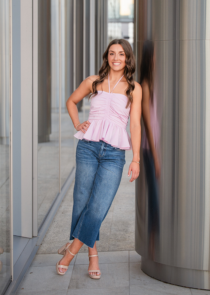 Macey's senior photos wearing a pink halter top and jeans leans casually against a reflective column. She's outside in an urban setting, exuding confidence.