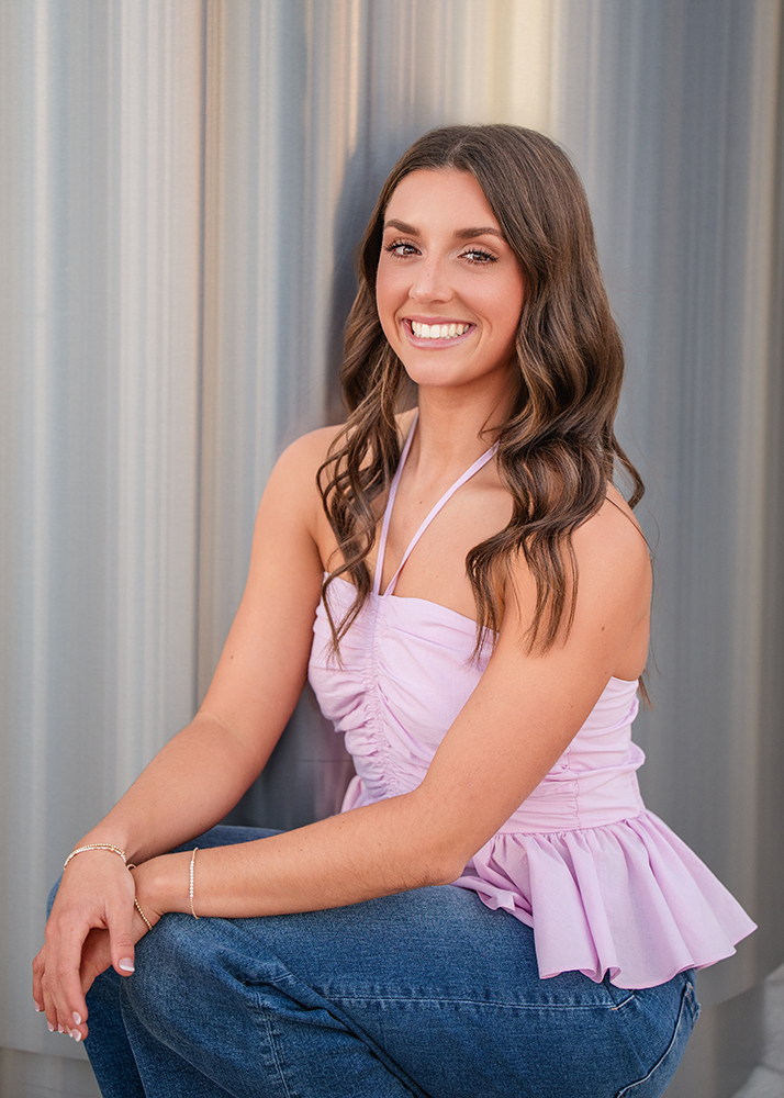 A smiling woman with long brown hair poses in a sleeveless pink top and blue jeans against a metallic background, exuding a cheerful and relaxed vibe.