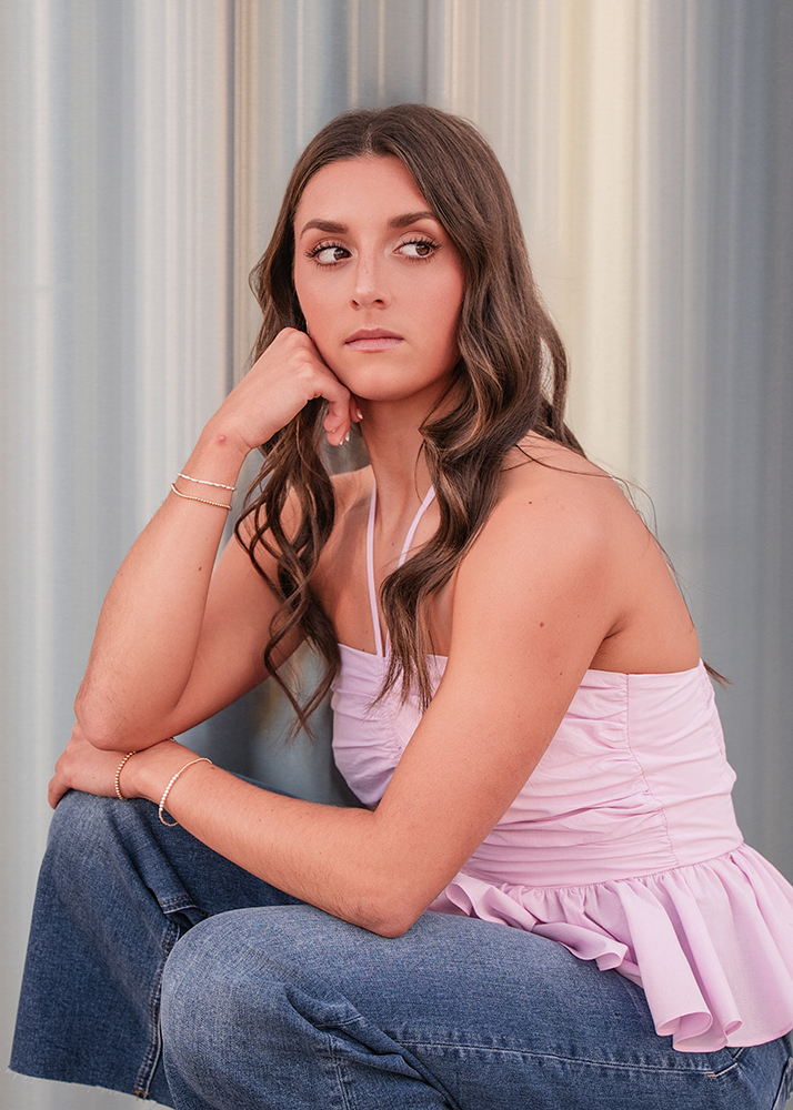 Young woman with long wavy hair gazes to the side thoughtfully. She wears a pink halter top and blue jeans against a soft gradient background.