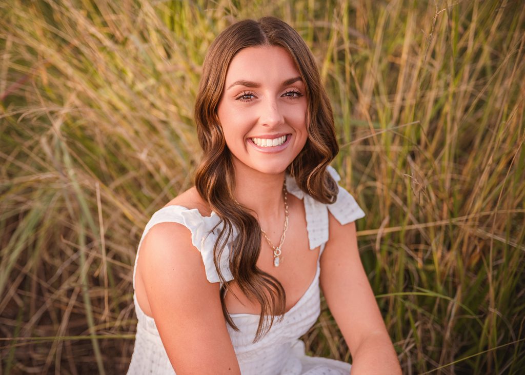 High school senior with long brown hair and a white dress smiles warmly, seated in front of tall grass. The setting is natural and serene.