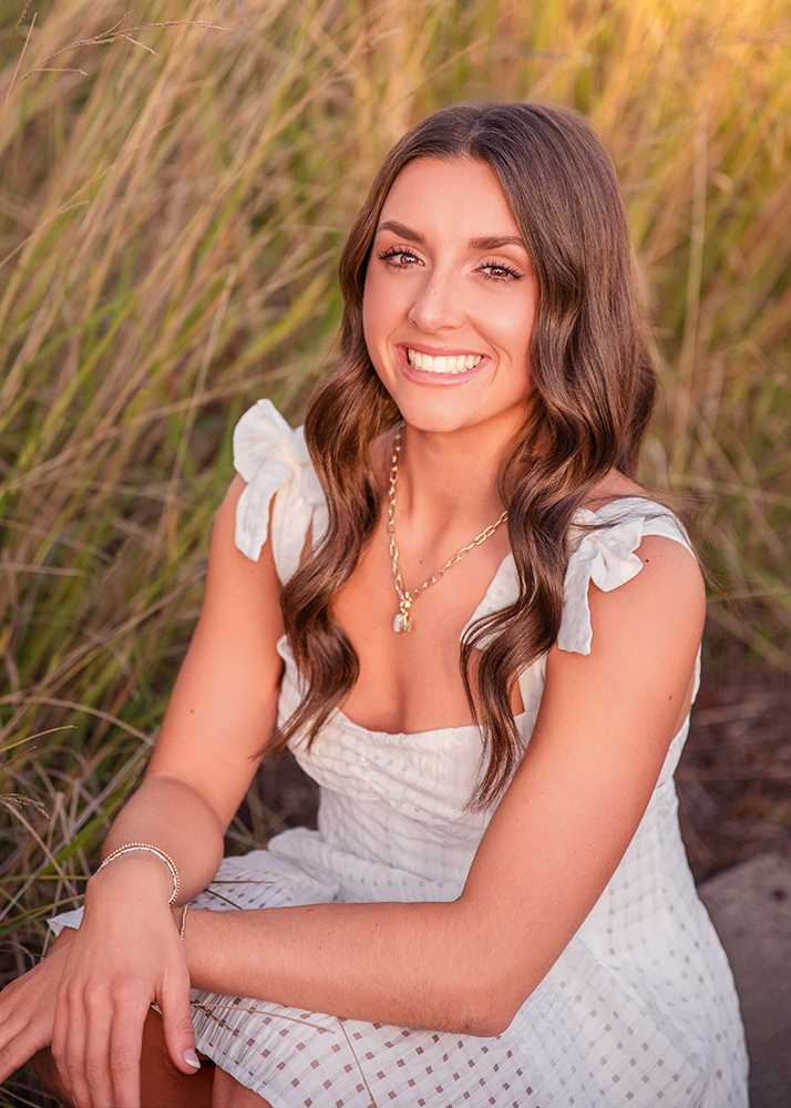 Macey sits in a field of tall grass, wearing a white dress with ruffled sleeves and a gold necklace for her senior photos. The scene conveys warmth and serenity.