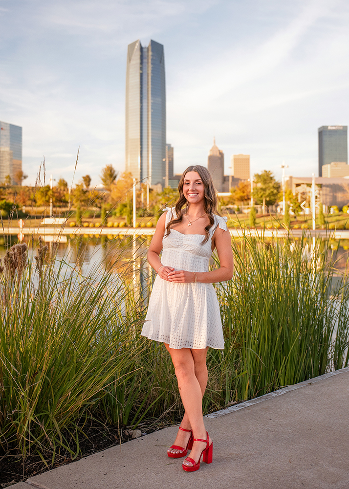 A high school senior in a white dress and red heels stands on a pathway near tall grass, smiling warmly. The backdrop shows the Oklahoma City cityscape behind her.