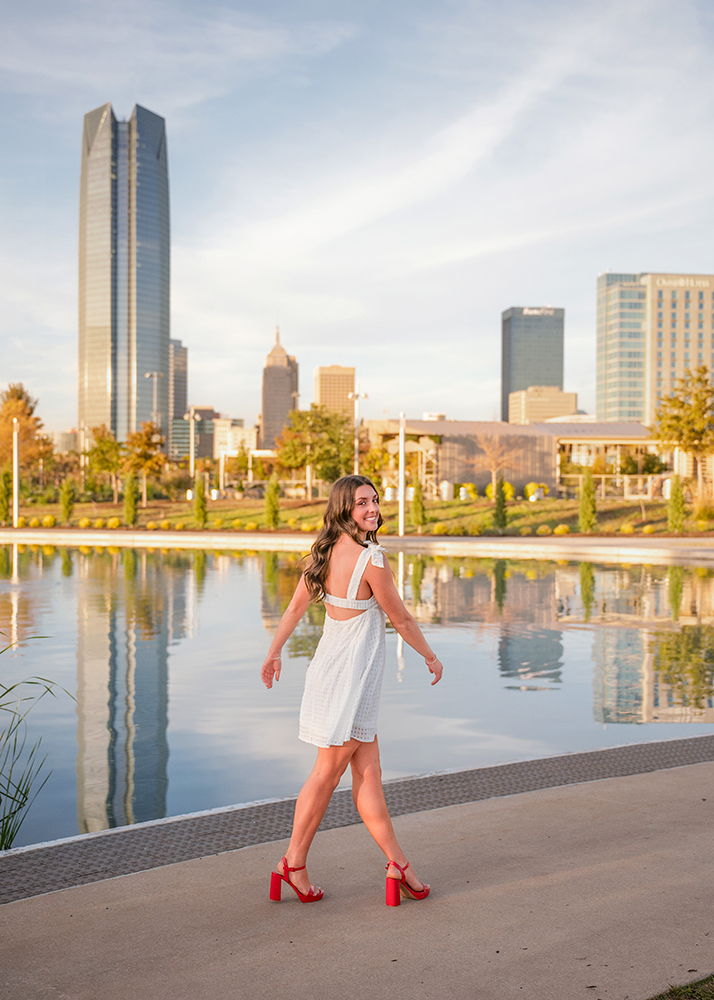 A woman in a white dress and red heels walks by a reflective water feature, with Oklahoma City skyline and trees in the background under a blue sky.