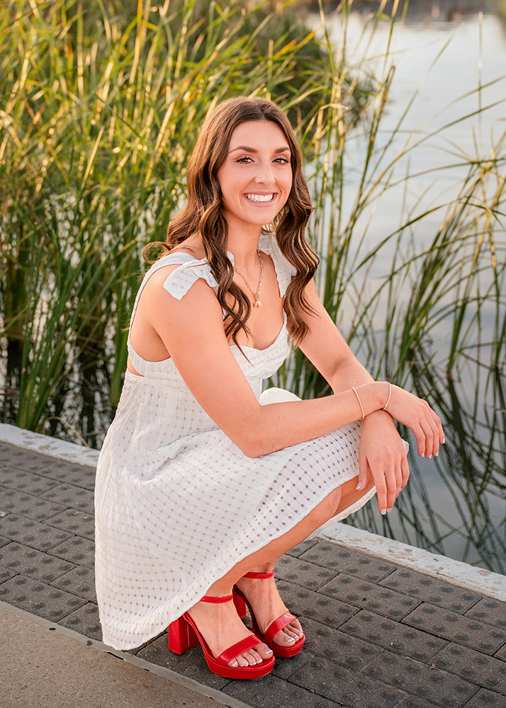 High school senior in a white dress and red heels smiles while crouching by a lakeside. Tall grass in the background; a serene and cheerful scene.