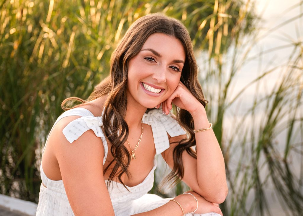 Macey's senior photos taken outdoors near green reeds. Warm, natural lighting enhances her joyful expression.