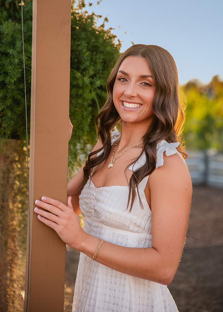 High school senior stands outdoors, smiling and leaning against a wooden post. The background features greenery bathed in warm, golden sunlight.