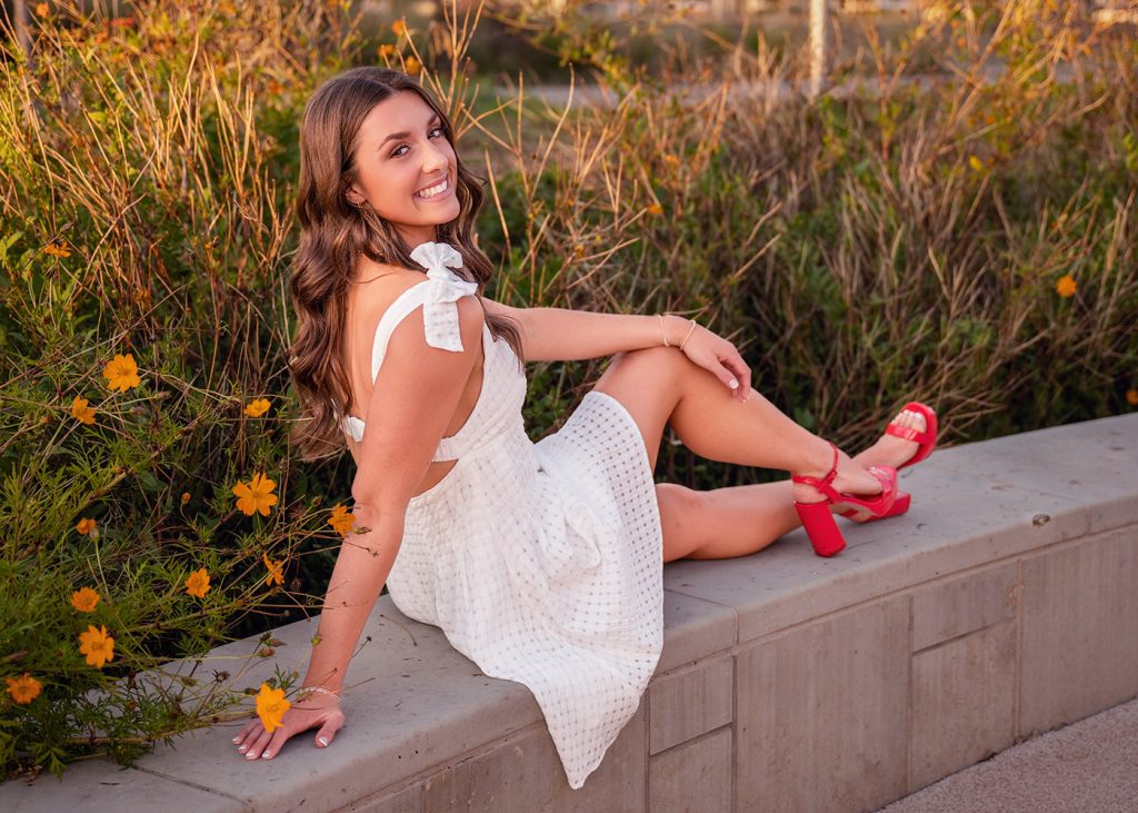 A senior girl in a white dress and red heels sits on a concrete ledge, smiling at the camera. She's surrounded by yellow flowers and green foliage.