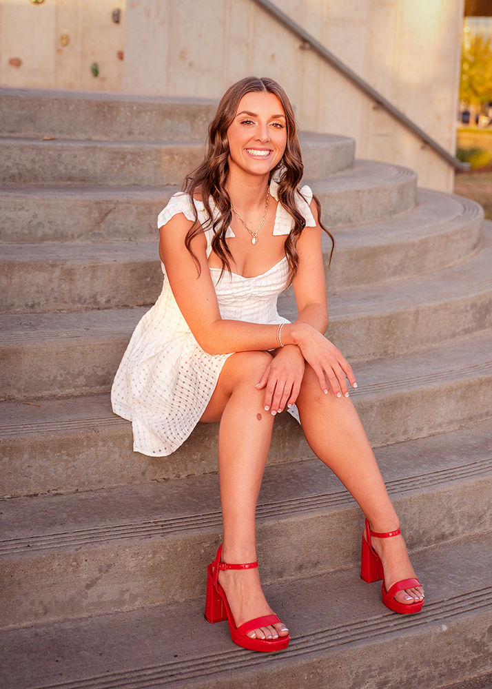 Macey takes her senior photos in a white dress sits on stone steps, smiling warmly. She wears red high-heeled sandals. The scene is bright and cheerful, with sunlight casting a soft glow.