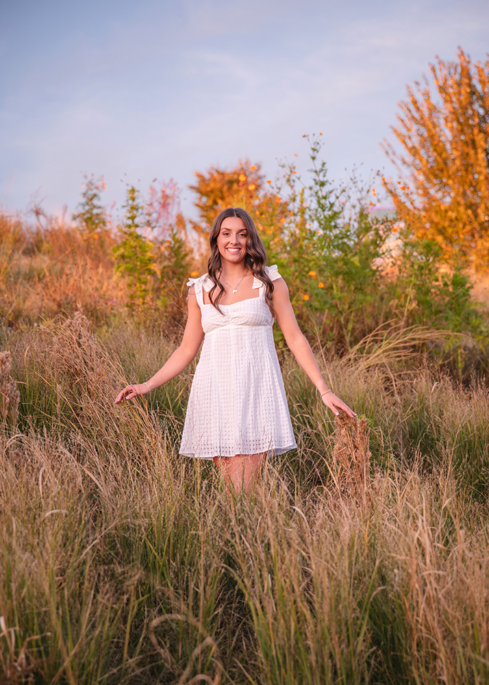 Smiling senior girl in a white dress stands in tall grasses, surrounded by vibrant autumn foliage. The scene is serene, with a warm, golden sunset glow.