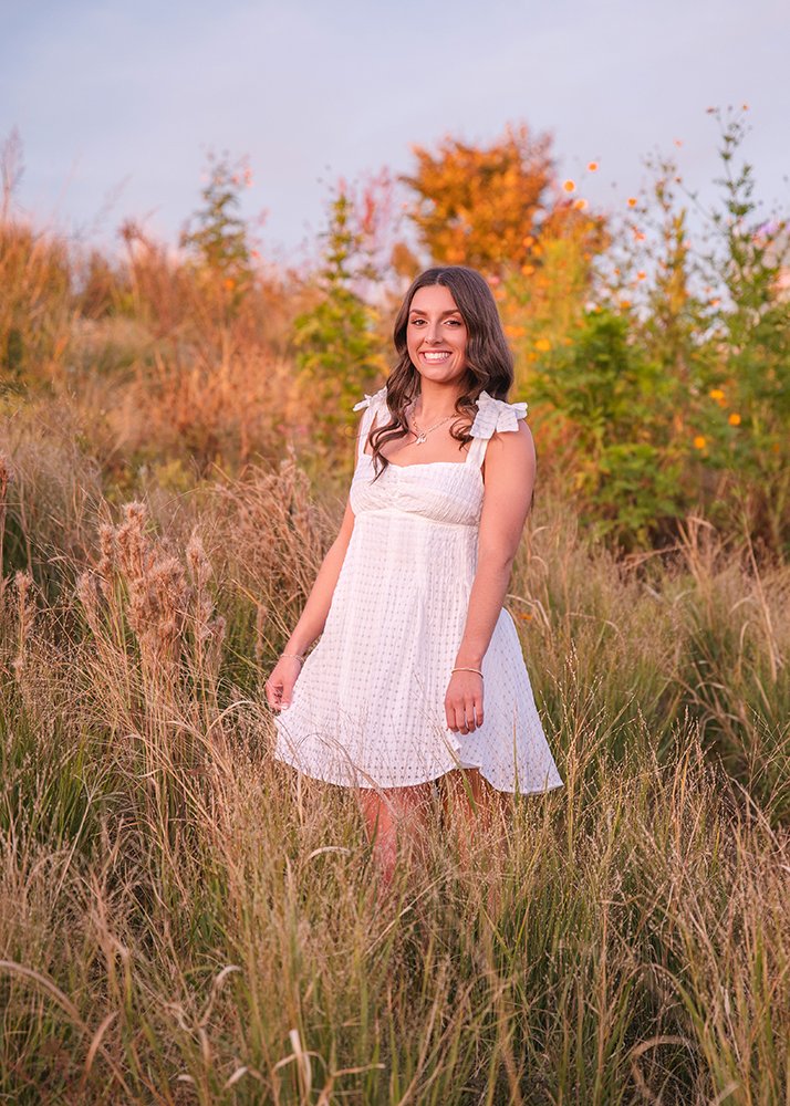 High school senior in a white dress stands smiling in a sunlit field of tall grass, with colorful foliage behind her, conveying a serene and joyful mood.