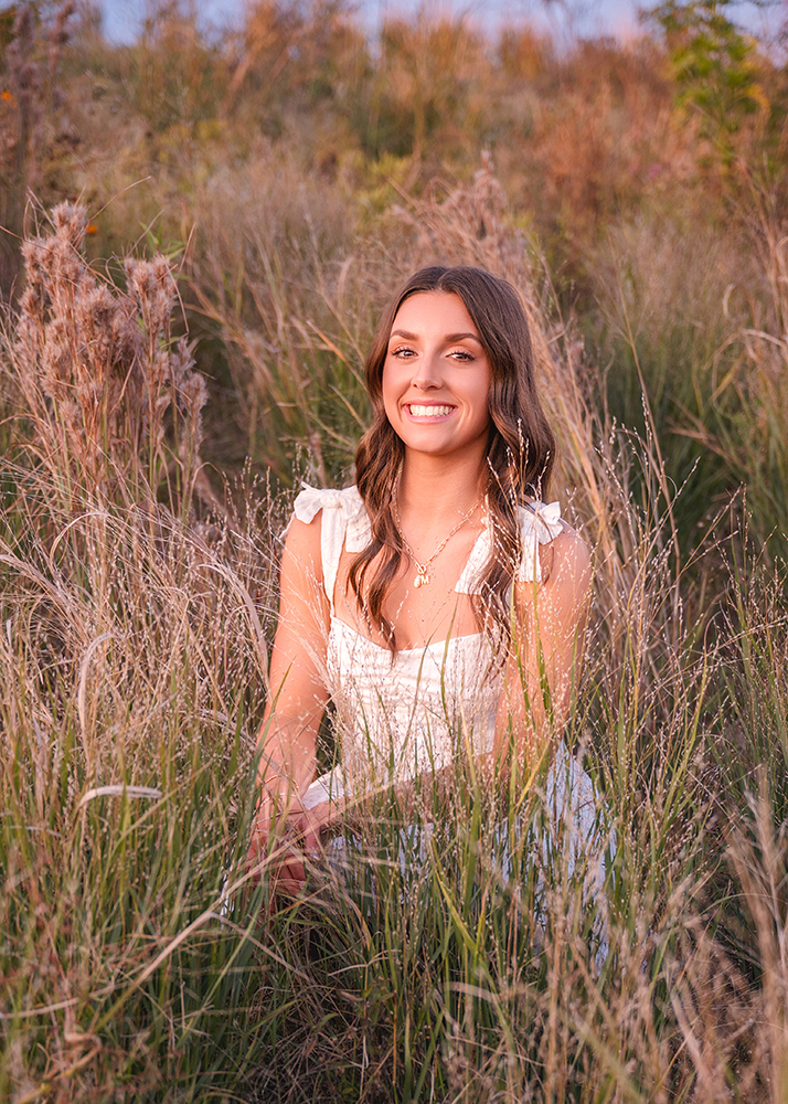 Senior girl in a white dress sits smiling amid tall, dry grass in a rustic field, evoking a serene and warm mood during sunset.