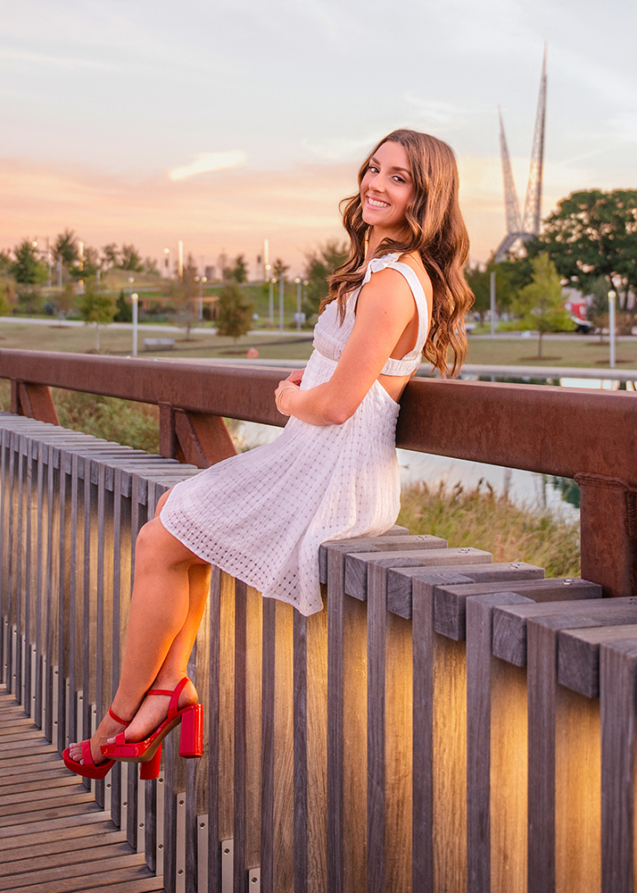A high school senior in a white dress and red heels sits smiling on a bridge railing at sunset. The background features Scissortail Park in downtown Oklahoma City.